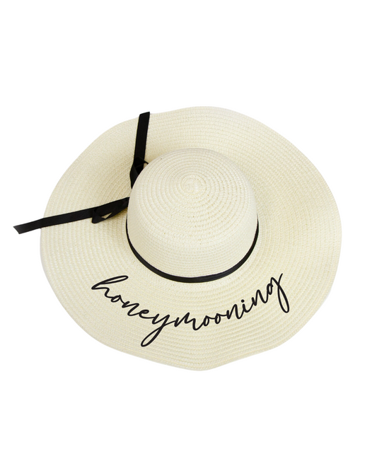 Flay lay on a white backdrop of an ivory floppy beach hat with the word 'honeymooning' in black cursive on the brim and black ribbon around the band.