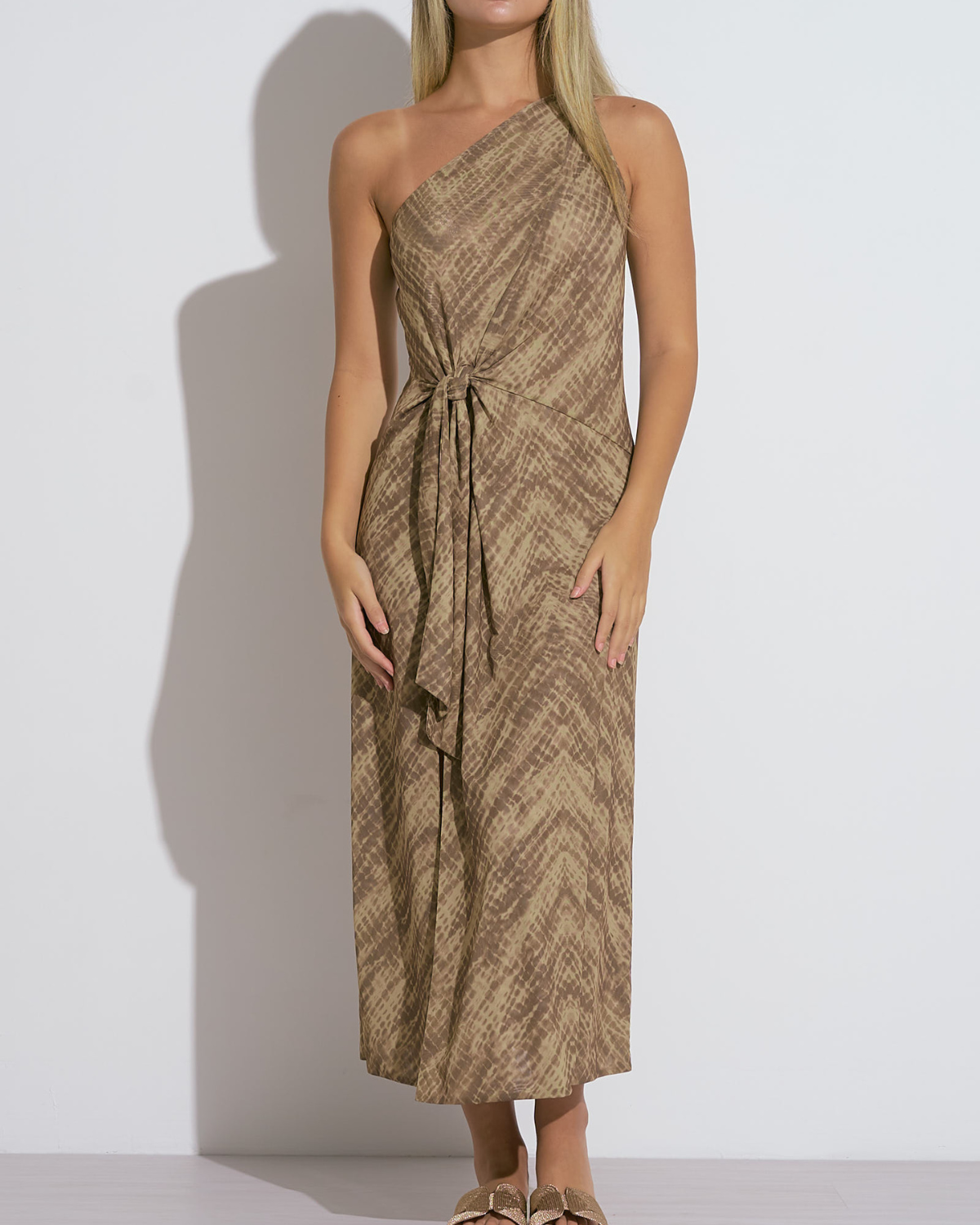A woman wearing a brown one-shoulder midi dress with a subtle chevron-style textured pattern. The dress features a waist tie detail that adds definition and a touch of elegance to the relaxed silhouette. She pairs the outfit with flat, gold-toned bow sandals, completing a laid-back yet polished warm-weather look.