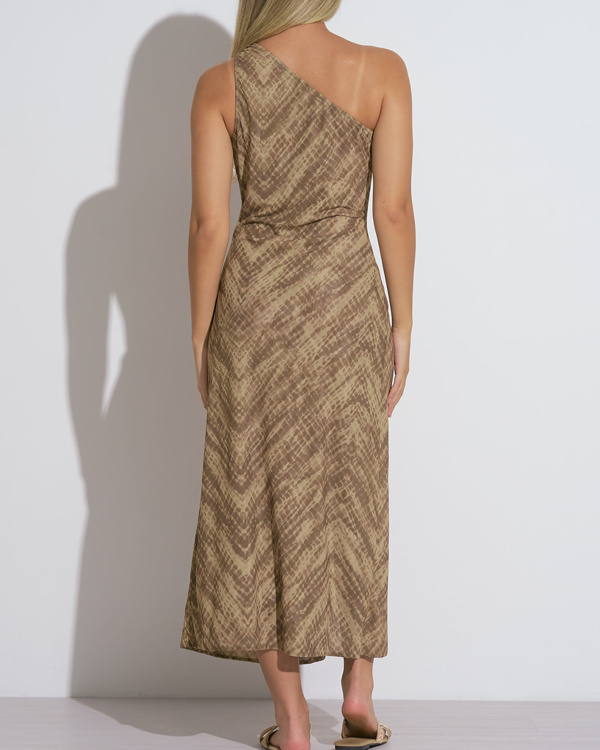 A woman wearing a brown one-shoulder midi dress with a subtle chevron-style textured pattern. The dress features a waist tie detail that adds definition and a touch of elegance to the relaxed silhouette. She pairs the outfit with flat, gold-toned bow sandals, completing a laid-back yet polished warm-weather look.