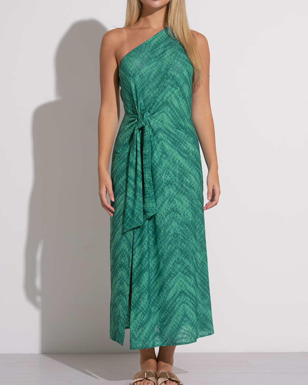 A woman wearing a green one-shoulder midi dress with a subtle textured pattern. The dress features a side knot at the waist, creating a gathered effect and a high front slit that adds movement and a hint of allure. She pairs the outfit with flat sandals featuring gold bow details, creating a chic and relaxed look suitable for warm weather or a resort setting.