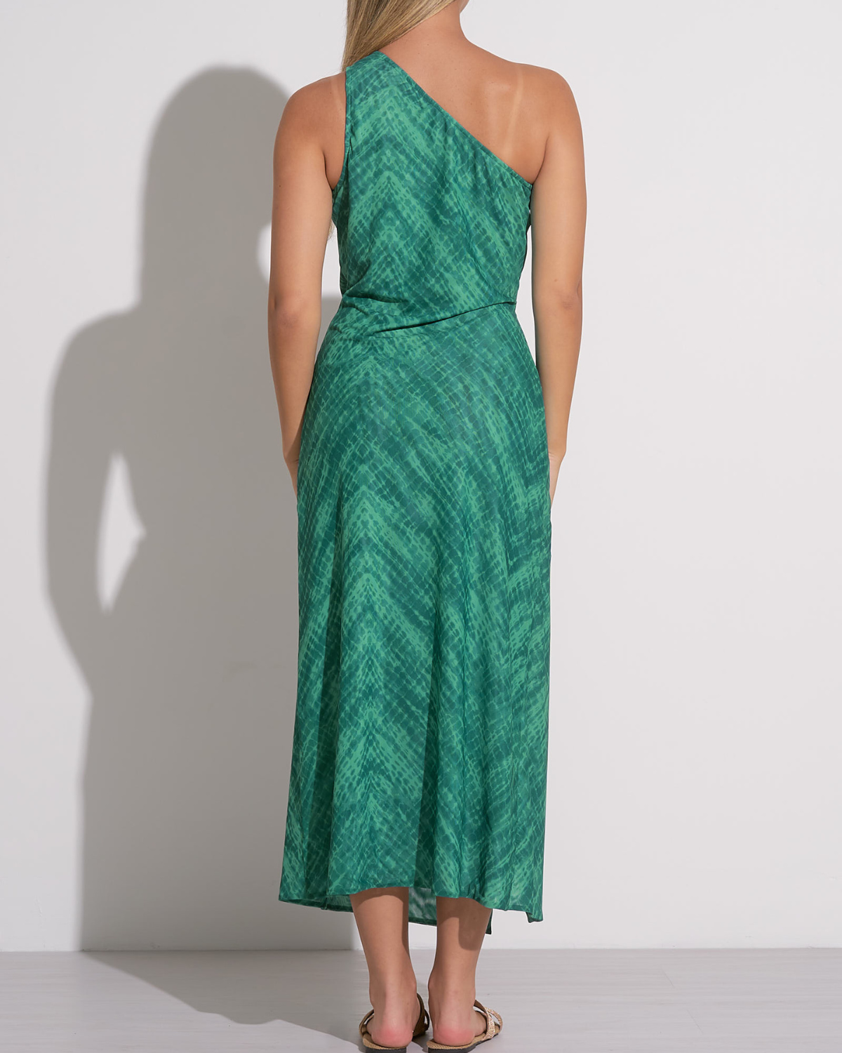 A woman wearing a green one-shoulder midi dress with a subtle textured pattern. The dress features a side knot at the waist, creating a gathered effect and a high front slit that adds movement and a hint of allure. She pairs the outfit with flat sandals featuring gold bow details, creating a chic and relaxed look suitable for warm weather or a resort setting.