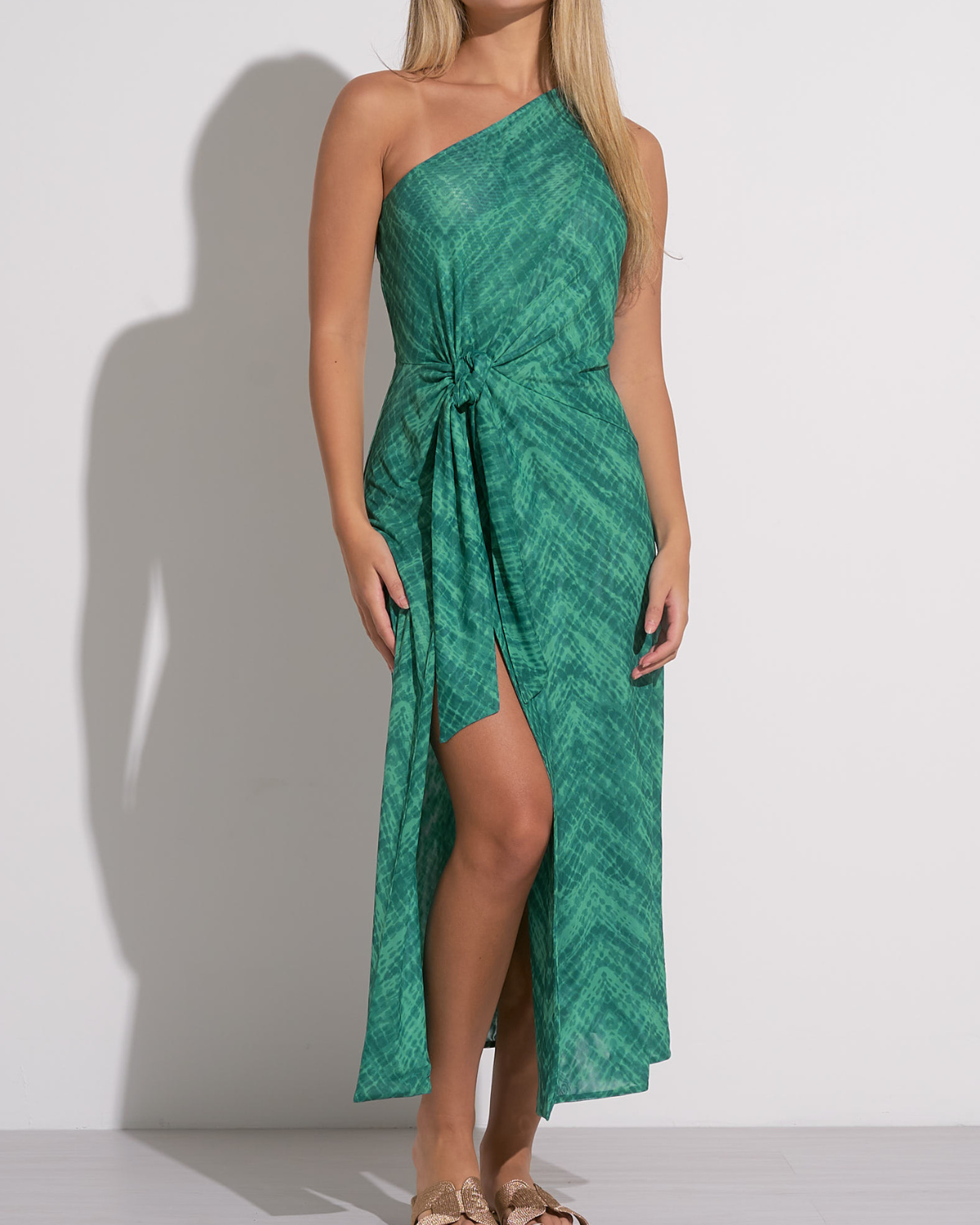 A woman wearing a green one-shoulder midi dress with a subtle textured pattern. The dress features a side knot at the waist, creating a gathered effect and a high front slit that adds movement and a hint of allure. She pairs the outfit with flat sandals featuring gold bow details, creating a chic and relaxed look suitable for warm weather or a resort setting.