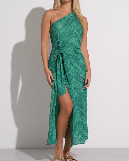 A woman wearing a green one-shoulder midi dress with a subtle textured pattern. The dress features a side knot at the waist, creating a gathered effect and a high front slit that adds movement and a hint of allure. She pairs the outfit with flat sandals featuring gold bow details, creating a chic and relaxed look suitable for warm weather or a resort setting.