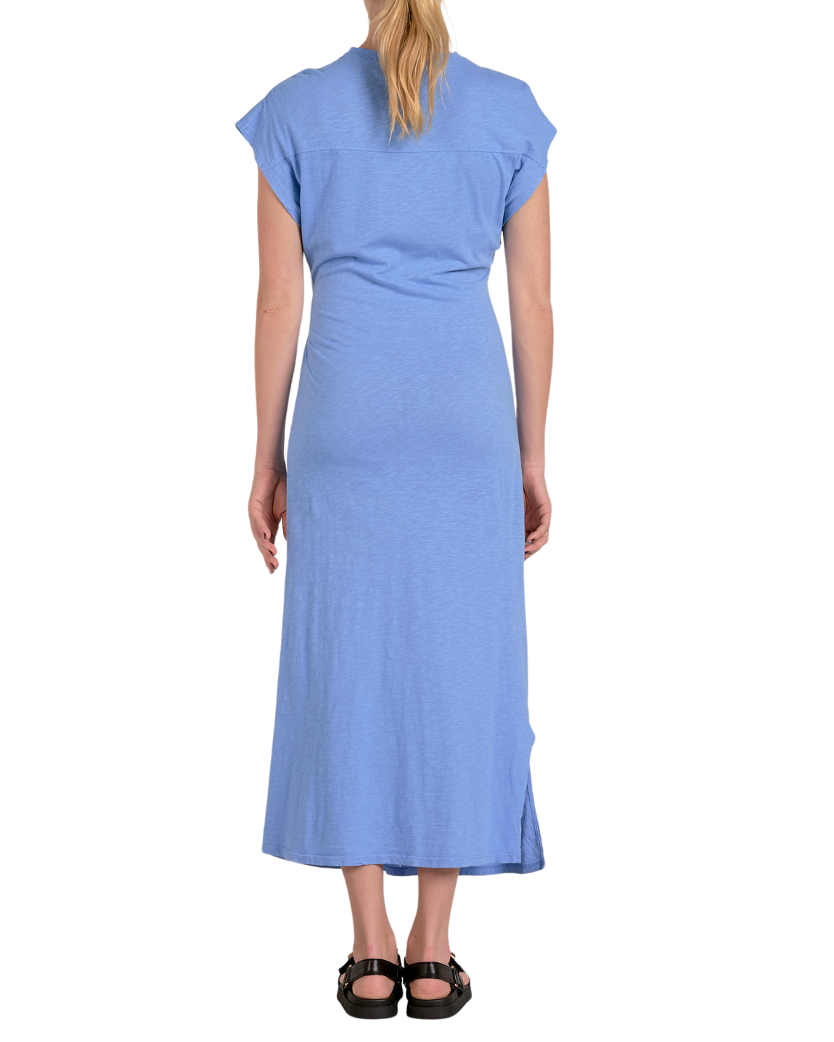 A woman wearing a light blue, short-sleeved midi dress made from a soft, jersey-like fabric. The dress features a crew neckline, a side tie detail at the waist creating a gathered effect, and a side slit for added movement. She completes the look with black buckle sandals and subtle stud earrings.