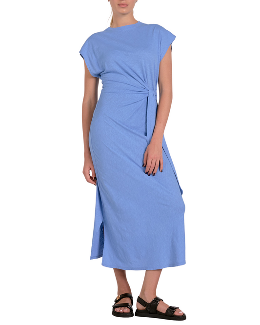 A woman wearing a light blue, short-sleeved midi dress made from a soft, jersey-like fabric. The dress features a crew neckline, a side tie detail at the waist creating a gathered effect, and a side slit for added movement. She completes the look with black buckle sandals and subtle stud earrings.