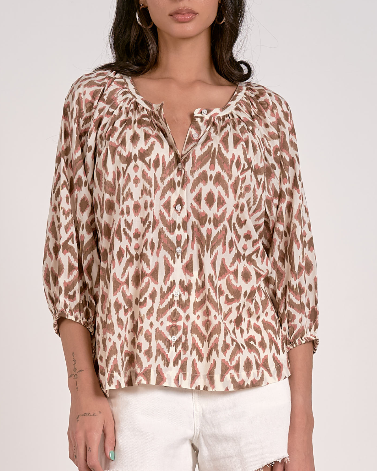 A woman wearing a loose-fitting blouse with a brown and pink abstract animal print pattern. The blouse features a scoop neckline with a partial button-down front, gathered detailing at the shoulders, and three-quarter length sleeves with elastic cuffs. She pairs it with white, high-waisted denim shorts with a raw hem, accessorized with hoop earrings and a natural makeup look.