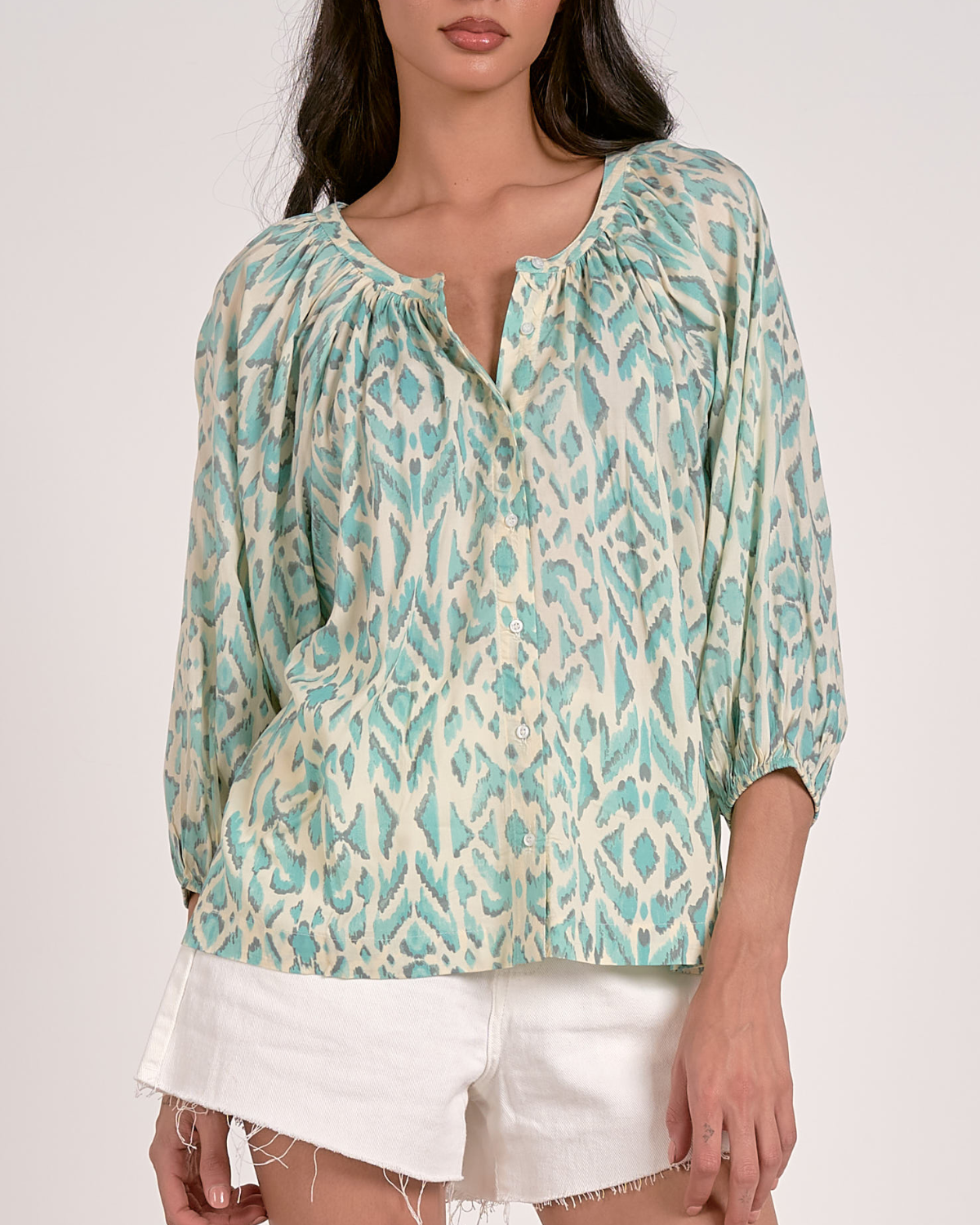 A woman wearing a loose-fitting blouse with a blue abstract animal print pattern. The blouse features a scoop neckline with a partial button-down front, gathered detailing at the shoulders, and three-quarter length sleeves with elastic cuffs. She pairs it with white, high-waisted denim shorts with a raw hem, accessorized with hoop earrings and a natural makeup look.