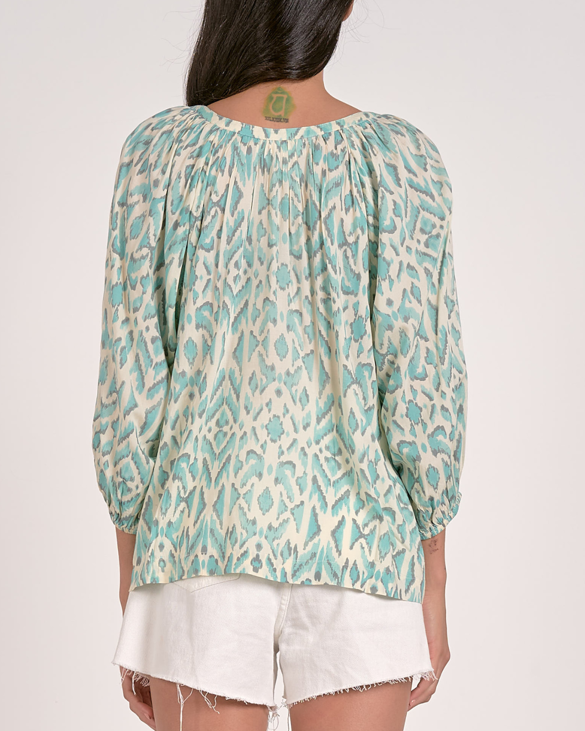 A woman wearing a loose-fitting blouse with a blue abstract animal print pattern. The blouse features a scoop neckline with a partial button-down front, gathered detailing at the shoulders, and three-quarter length sleeves with elastic cuffs. She pairs it with white, high-waisted denim shorts with a raw hem, accessorized with hoop earrings and a natural makeup look.