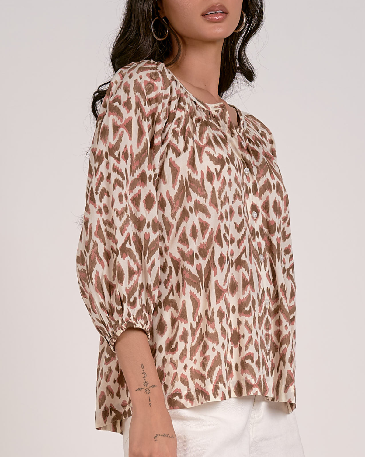 A woman wearing a loose-fitting blouse with a brown and pink abstract animal print pattern. The blouse features a scoop neckline with a partial button-down front, gathered detailing at the shoulders, and three-quarter length sleeves with elastic cuffs. She pairs it with white, high-waisted denim shorts with a raw hem, accessorized with hoop earrings and a natural makeup look.