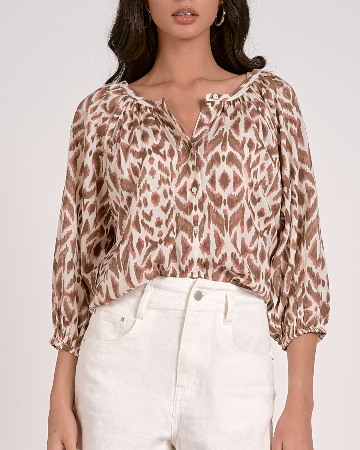 A woman wearing a loose-fitting blouse with a brown and pink abstract animal print pattern. The blouse features a scoop neckline with a partial button-down front, gathered detailing at the shoulders, and three-quarter length sleeves with elastic cuffs. She pairs it with white, high-waisted denim shorts with a raw hem, accessorized with hoop earrings and a natural makeup look.