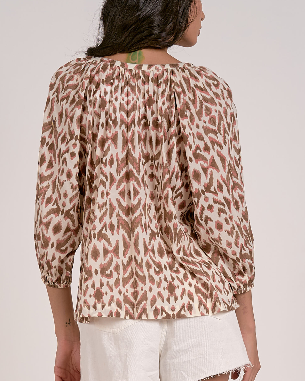 A woman wearing a loose-fitting blouse with a brown and pink abstract animal print pattern. The blouse features a scoop neckline with a partial button-down front, gathered detailing at the shoulders, and three-quarter length sleeves with elastic cuffs. She pairs it with white, high-waisted denim shorts with a raw hem, accessorized with hoop earrings and a natural makeup look.