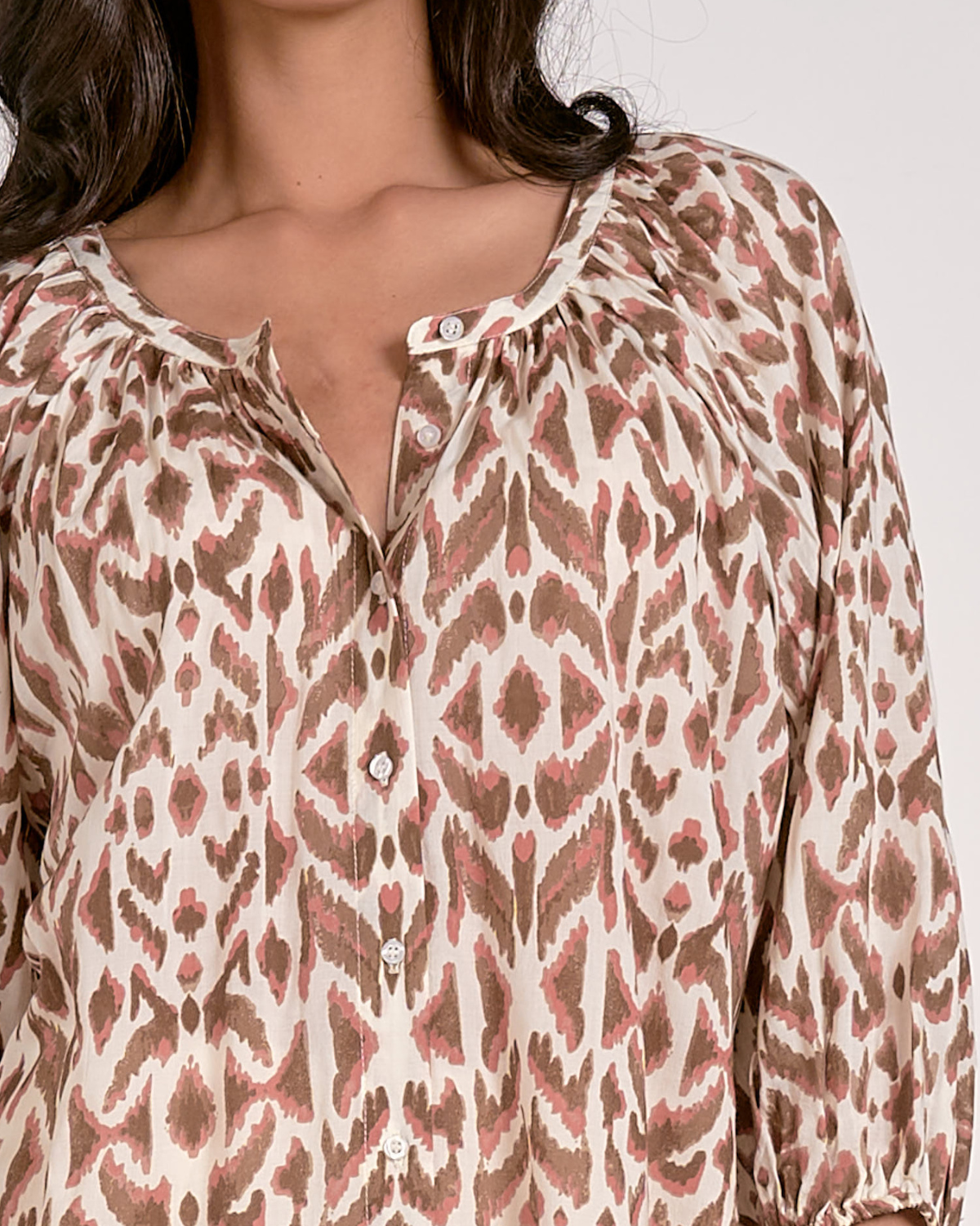 A woman wearing a loose-fitting blouse with a brown and pink abstract animal print pattern. The blouse features a scoop neckline with a partial button-down front, gathered detailing at the shoulders, and three-quarter length sleeves with elastic cuffs. She pairs it with white, high-waisted denim shorts with a raw hem, accessorized with hoop earrings and a natural makeup look.