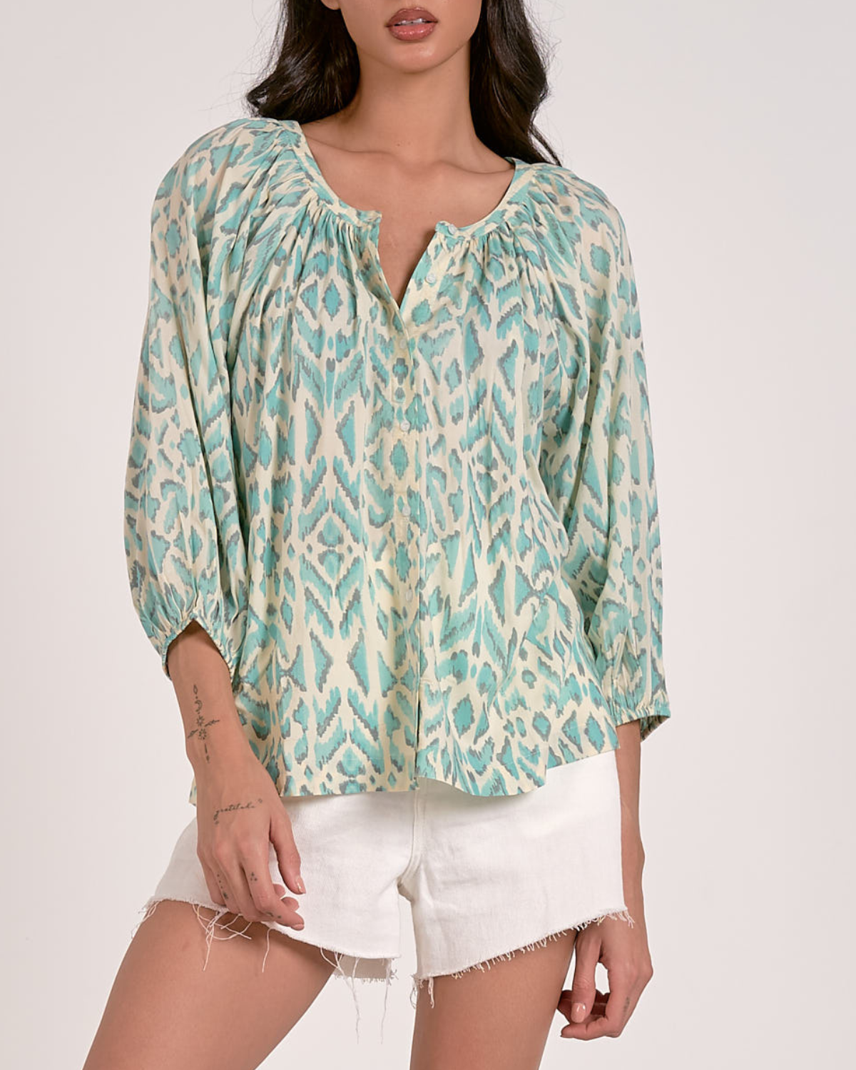 A woman wearing a loose-fitting blouse with a blue abstract animal print pattern. The blouse features a scoop neckline with a partial button-down front, gathered detailing at the shoulders, and three-quarter length sleeves with elastic cuffs. She pairs it with white, high-waisted denim shorts with a raw hem, accessorized with hoop earrings and a natural makeup look.