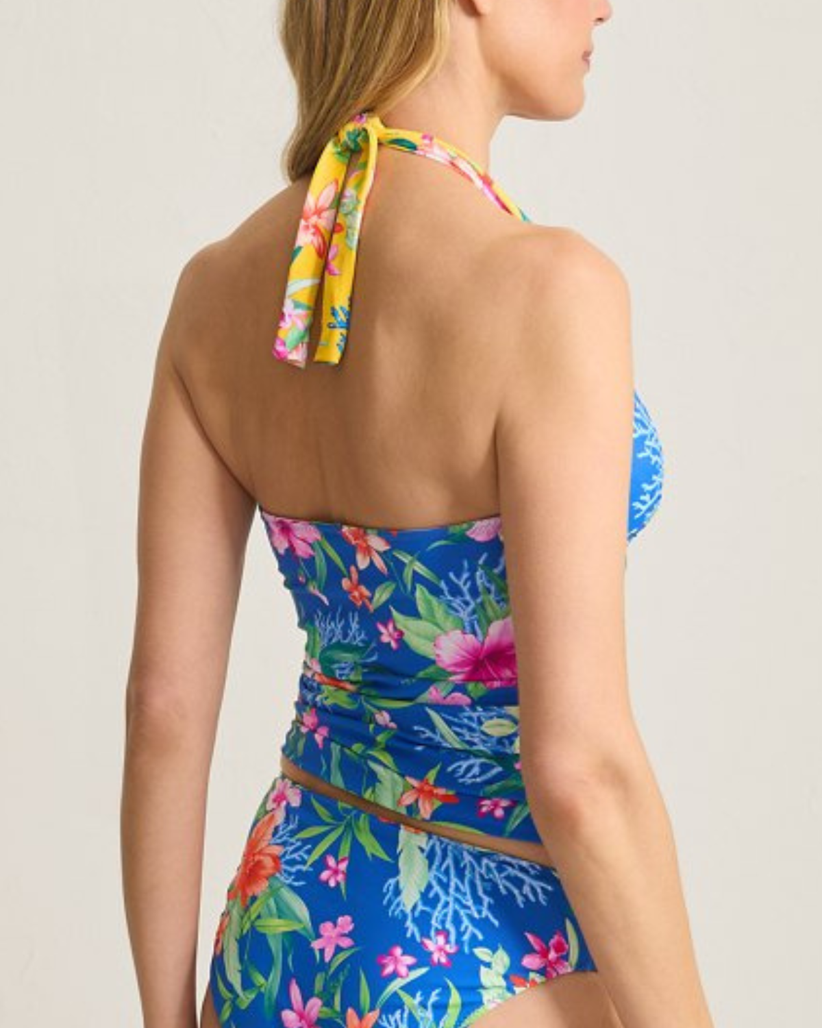 A woman wearing a tropical floral halter tankini top in a vibrant blue color. The top features a ruched design along the sides and bust for a flattering fit, with pink, orange, and green floral details and coral reef accents. The neckline is a deep V with adjustable halter straps.