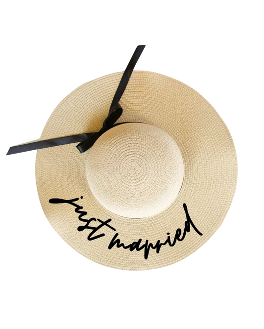 Flat lay on a white backdrop of a light tan floppy beach hat with the words 'Just Married' in a black cursive font on the brim and a black ribbon band.