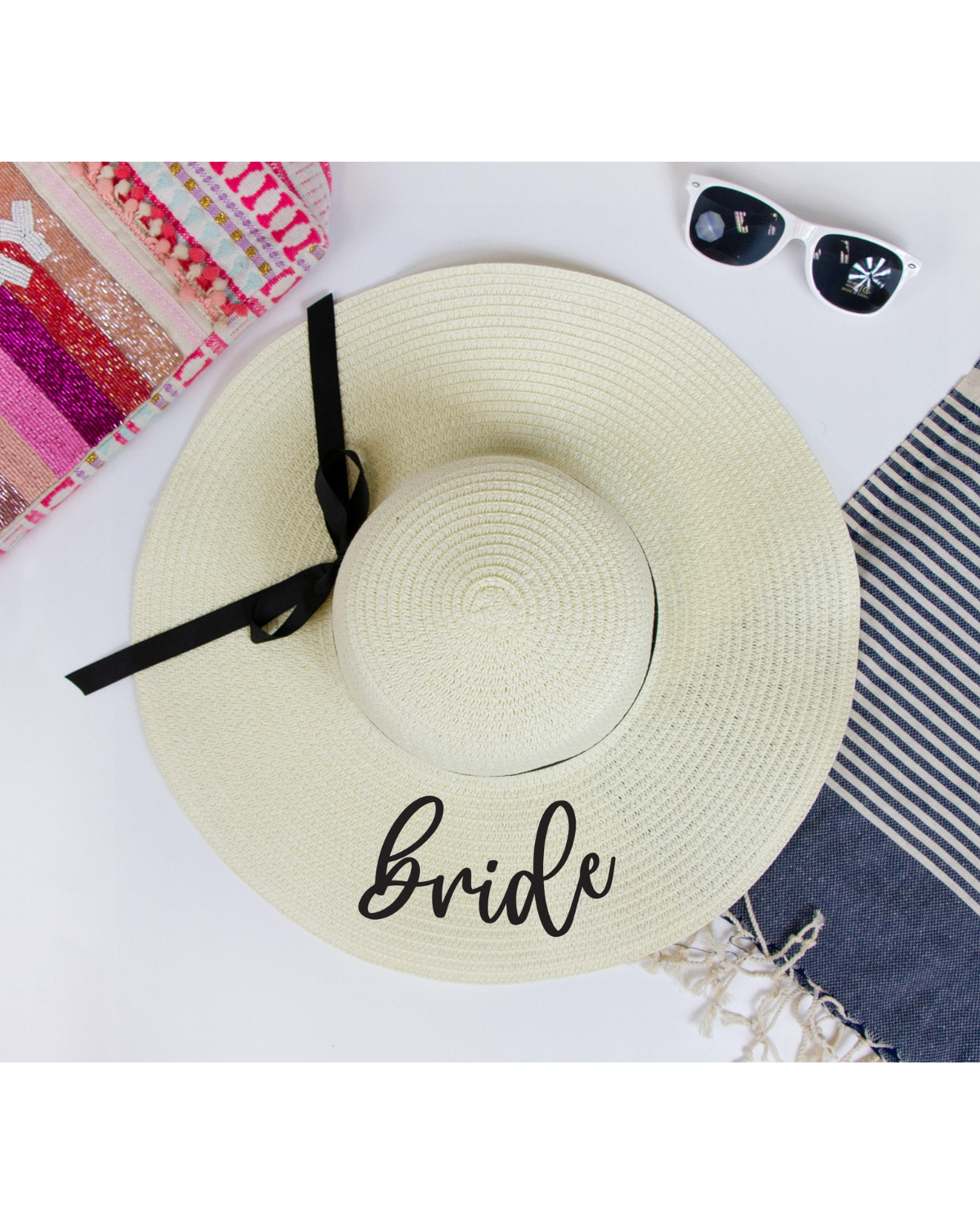 Flat lay on a white backdrop wearing ivory straw floppy hat with the word 'Bride' on the brim with a black ribbon band. Beach accessories surrounding the hat.