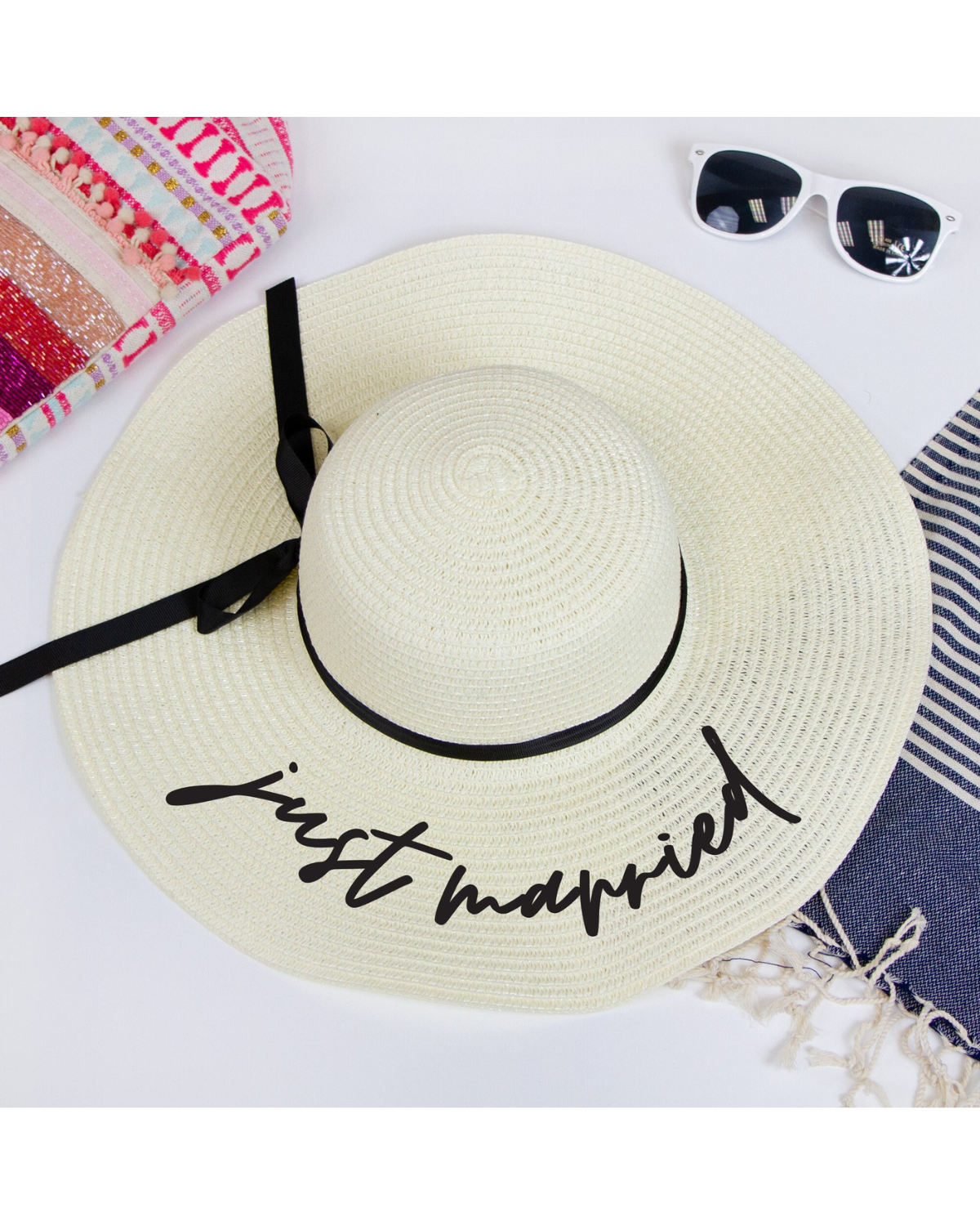 Flat lay on a white backdrop wearing ivory straw floppy hat with the words 'Just Married' on the brim and a black ribbon band. Beach accessories surrounding the hat. 