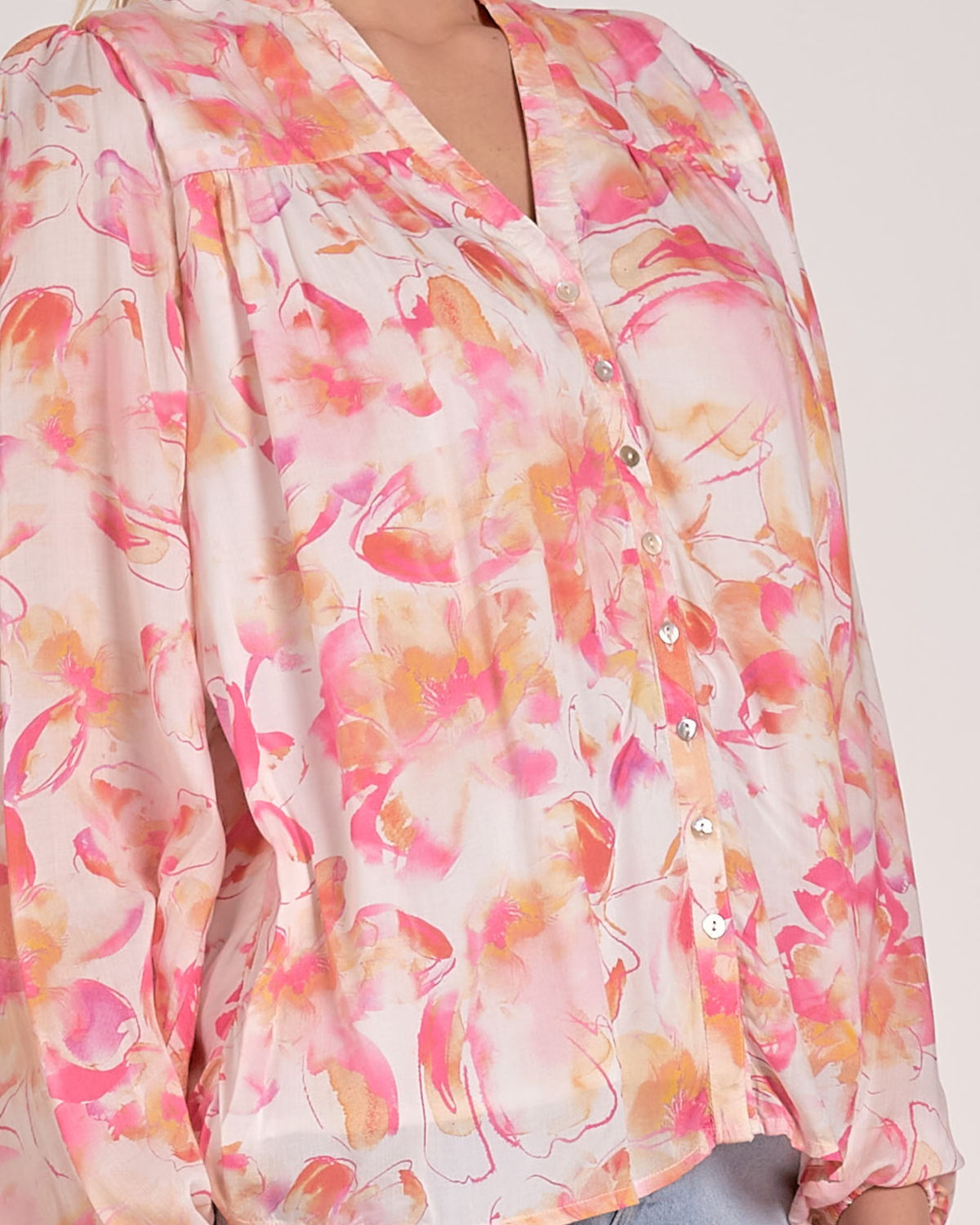 A woman wearing a lightweight, long-sleeved blouse with a vibrant watercolor floral print in shades of pink, orange, and yellow on a light background. The blouse features a V-neckline with a button-down front and a relaxed, flowy fit. She pairs it with light-wash, frayed denim shorts and simple stud earrings.