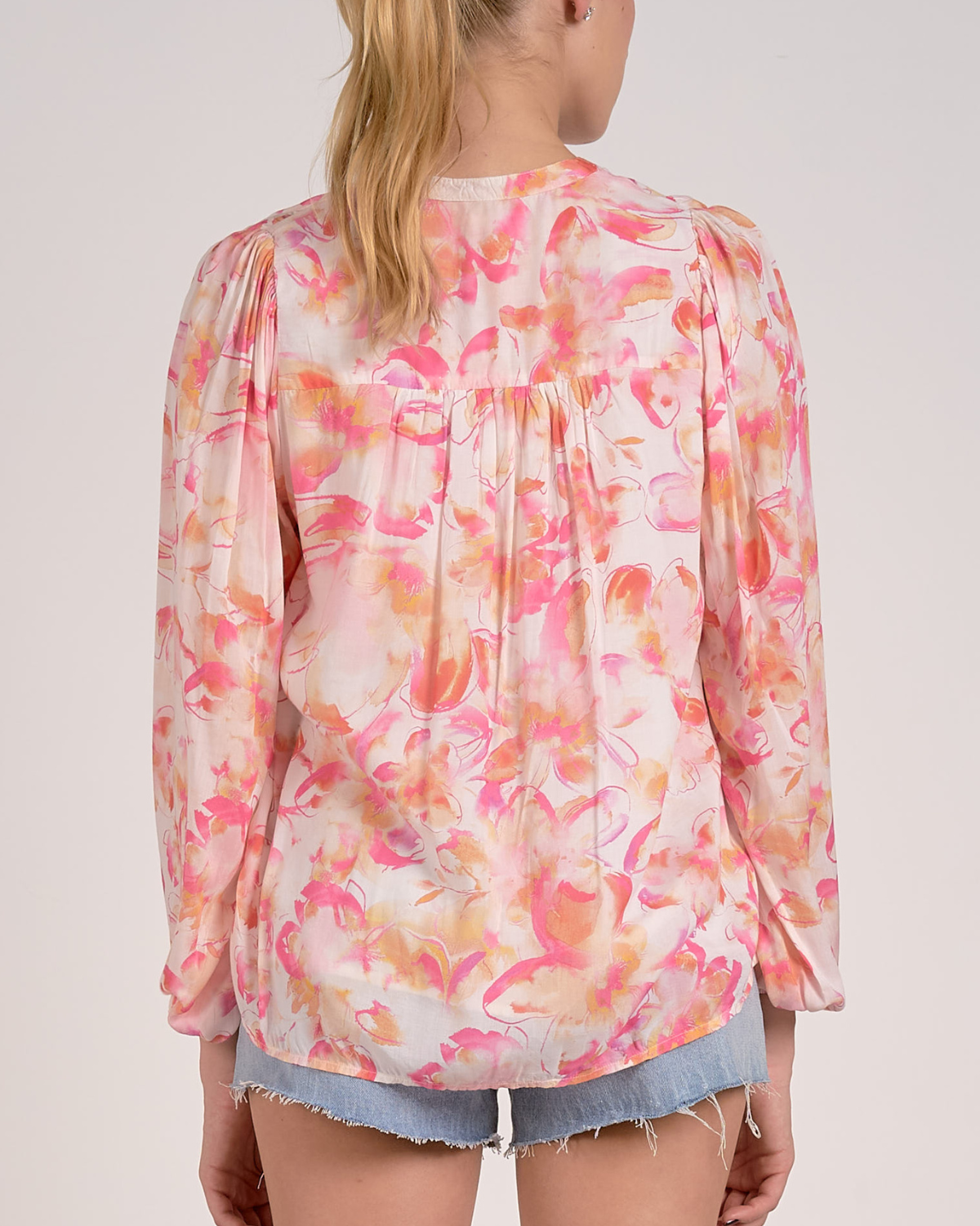A woman wearing a lightweight, long-sleeved blouse with a vibrant watercolor floral print in shades of pink, orange, and yellow on a light background. The blouse features a V-neckline with a button-down front and a relaxed, flowy fit. She pairs it with light-wash, frayed denim shorts and simple stud earrings.