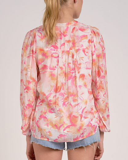 A woman wearing a lightweight, long-sleeved blouse with a vibrant watercolor floral print in shades of pink, orange, and yellow on a light background. The blouse features a V-neckline with a button-down front and a relaxed, flowy fit. She pairs it with light-wash, frayed denim shorts and simple stud earrings.