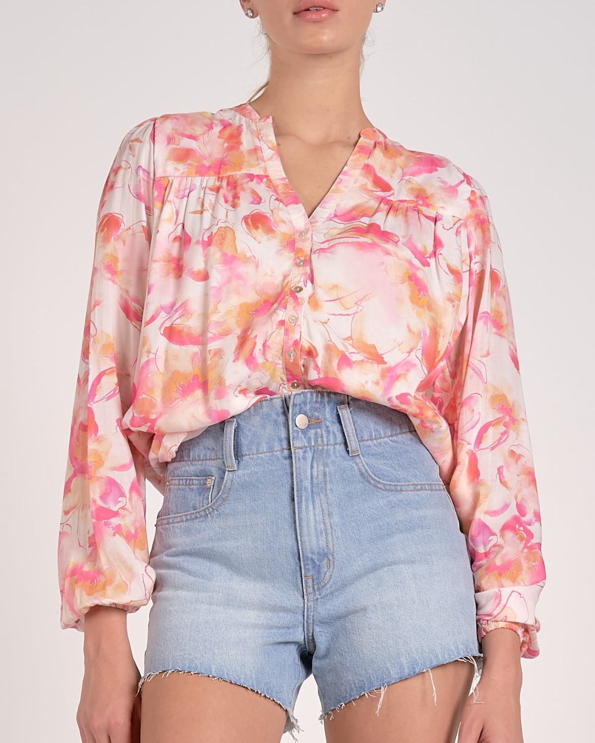 A woman wearing a lightweight, long-sleeved blouse with a vibrant watercolor floral print in shades of pink, orange, and yellow on a light background. The blouse features a V-neckline with a button-down front and a relaxed, flowy fit. She pairs it with light-wash, frayed denim shorts and simple stud earrings.