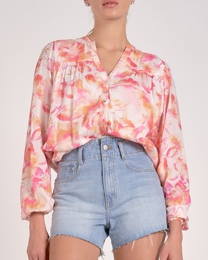 A woman wearing a lightweight, long-sleeved blouse with a vibrant watercolor floral print in shades of pink, orange, and yellow on a light background. The blouse features a V-neckline with a button-down front and a relaxed, flowy fit. She pairs it with light-wash, frayed denim shorts and simple stud earrings.