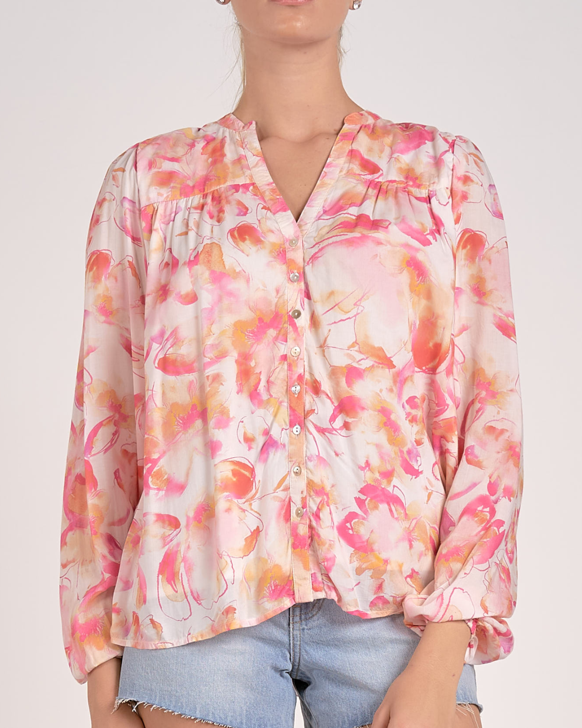 A woman wearing a lightweight, long-sleeved blouse with a vibrant watercolor floral print in shades of pink, orange, and yellow on a light background. The blouse features a V-neckline with a button-down front and a relaxed, flowy fit. She pairs it with light-wash, frayed denim shorts and simple stud earrings.