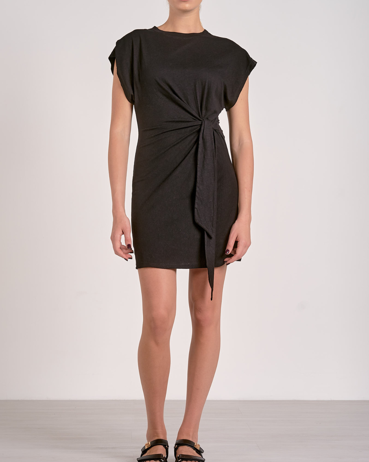 A woman wearing a black, short-sleeved mini dress made from a soft, stretch fabric. The dress features a round neckline and a stylish side knot detail at the waist, creating a flattering gathered effect. The hem is asymmetrical due to the tie design, adding a chic touch to the simple silhouette.