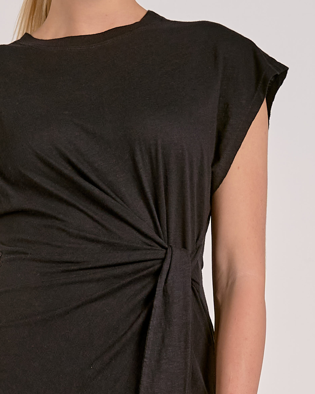 A woman wearing a black, short-sleeved mini dress made from a soft, stretch fabric. The dress features a round neckline and a stylish side knot detail at the waist, creating a flattering gathered effect. The hem is asymmetrical due to the tie design, adding a chic touch to the simple silhouette.
