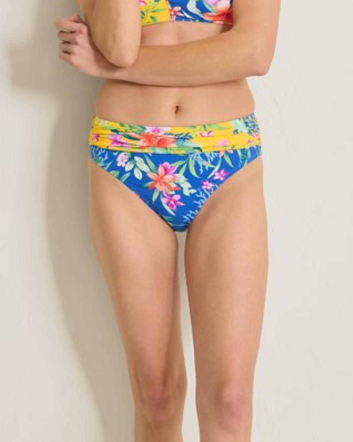 A woman wearing a tropical-print bikini bottom featuring a vibrant mix of floral patterns in pink, orange, and green on a bold blue and yellow background. The mid-rise bottom includes a wide, ruched waistband in yellow with floral accents, offering a flattering and comfortable fit.