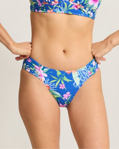 A reversible bikini bottom shown in two views. One side features a tropical floral print on a vibrant blue background, while the reverse side displays the same floral pattern on a bright yellow background. Both sides have a mid-rise fit with ruched detailing at the hips for a flattering silhouette. The design offers versatility and style in a sustainable swimwear option