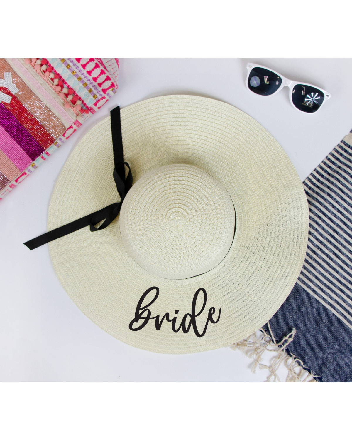 Flat lay on a white backdrop wearing ivory straw floppy hat with the word 'Bride' on the brim with a black ribbon band. Beach accessories surrounding the hat. 