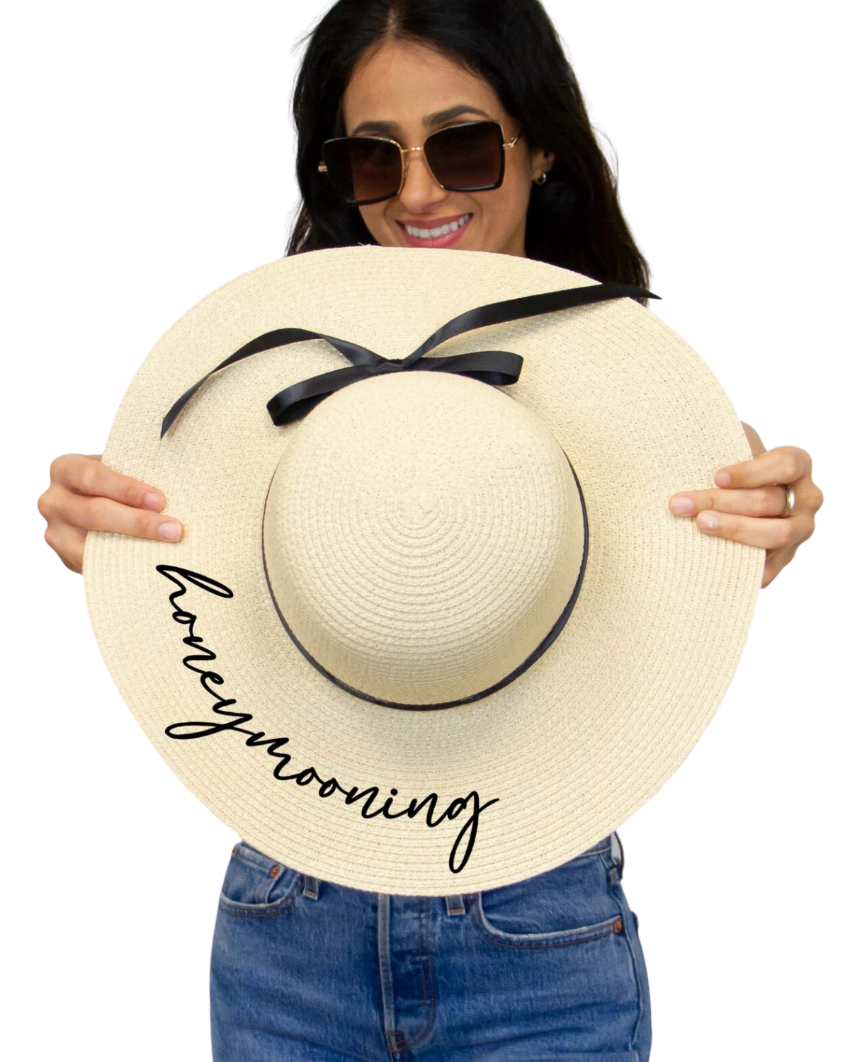 Model on a white backdrop holding an ivory floppy beach hat with the word 'honeymooning' in black cursive on the brim and black ribbon around the band.