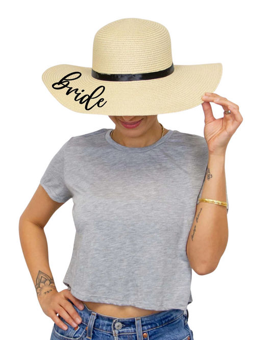 Model on a white backdrop wearing light tan straw floppy hat with the word 'Bride' on the brim and a black ribbon band.