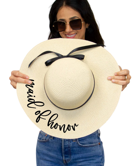 Model on a white backdrop holding an ivory floppy beach hat with the phrase 'Maid of Honor' in black cursive on the brim and black ribbon around the band.