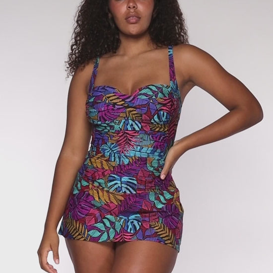 Model rotating 360 degrees in a swim dress with a shirred midsection in a navy, yellow, green and pink palm frond print.
