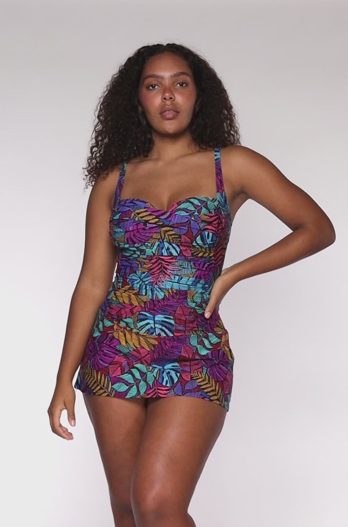 Model rotating 360 degrees in a swim dress with a shirred midsection in a navy, yellow, green and pink palm frond print.