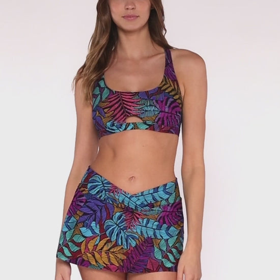 Model rotating 360 degrees wearing a v front swim skirt in a pink, purple, blue and yellow palm frond print.